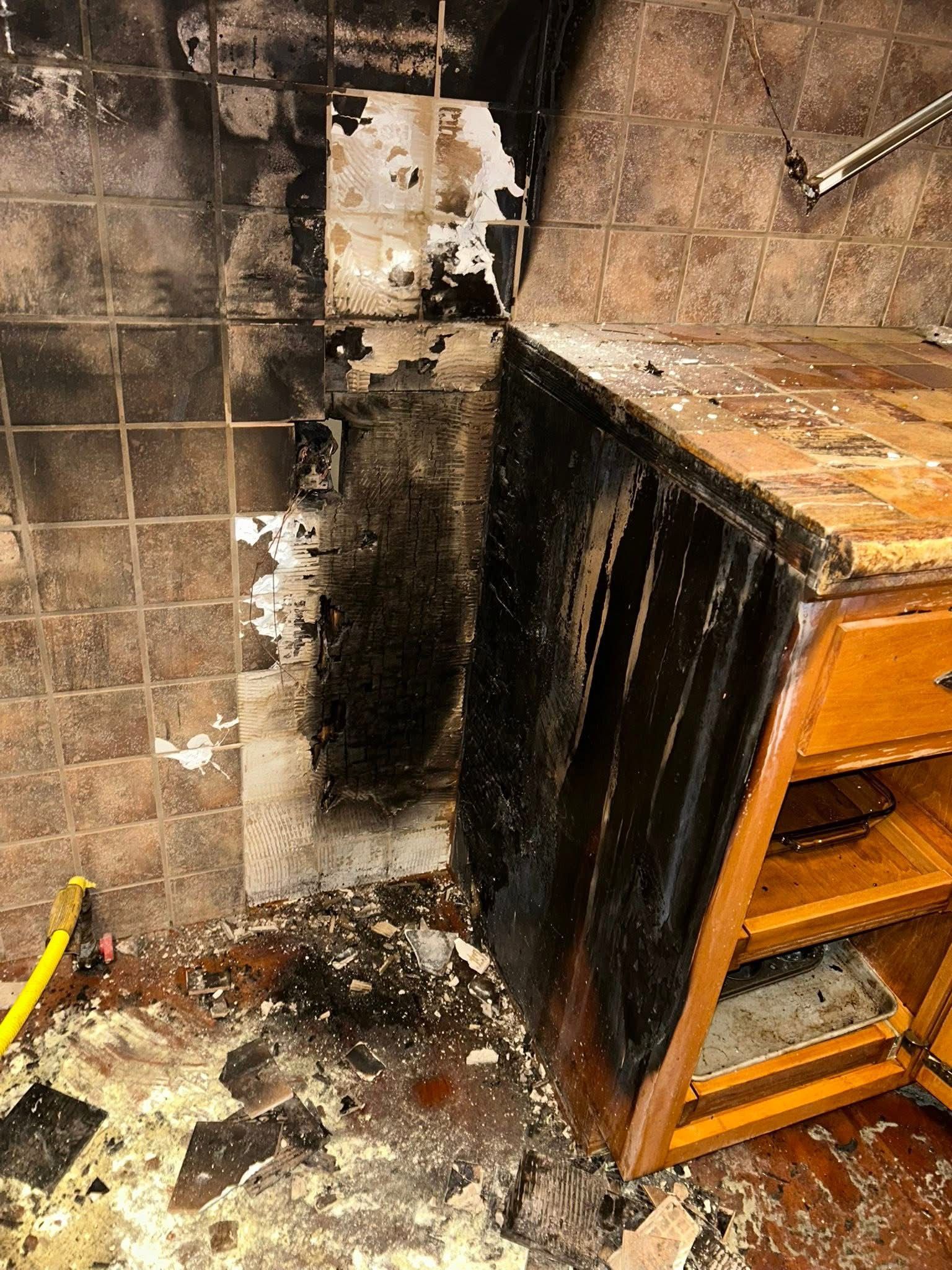 A charred corner of a kitchen with soot-stained tiles, burnt wooden cabinetry, and debris scattered across the floor.