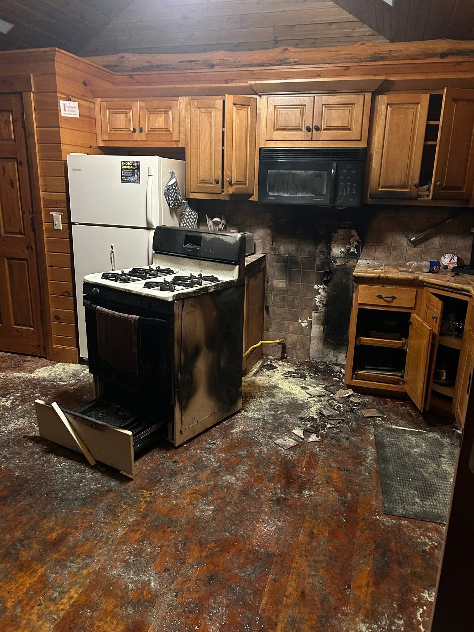 A kitchen in a log cabin with extensive fire damage, including a scorched stove, charred walls, and debris on the floor.