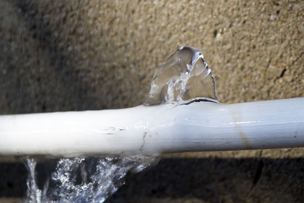 A White Pvc Pipe Bursts, Spraying Water Against a Rough Beige Wall 	— Ian Stanley Plumbing In Avoca, QLD