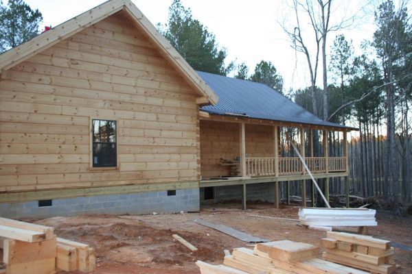A wooden house under construction with a covered porch and surrounding trees.