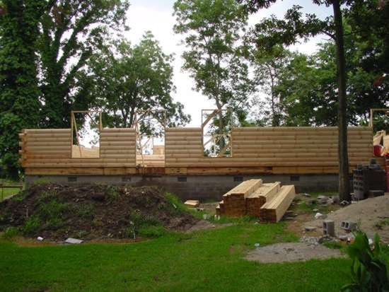Log cabin frame under construction on a concrete foundation, surrounded by green grass and trees.