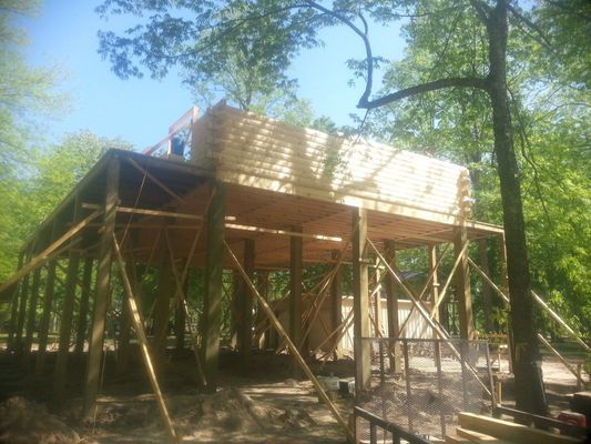 Wooden structure under construction in a wooded area; lumber stacked on top.