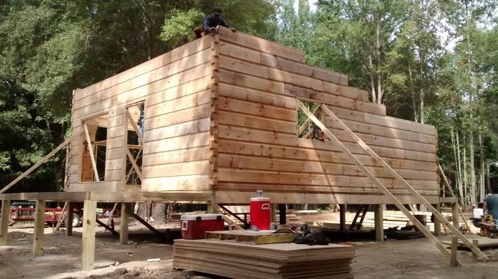 A log cabin being built on a raised wooden platform in a wooded area.