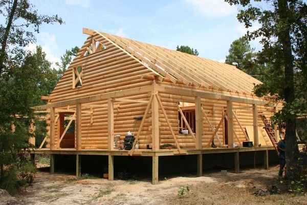 Wooden log cabin under construction, with exposed frame and unfinished roof, in a wooded area.