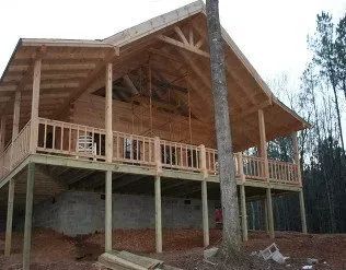 Wooden cabin under construction with a wraparound porch supported by wooden beams, trees in the background.