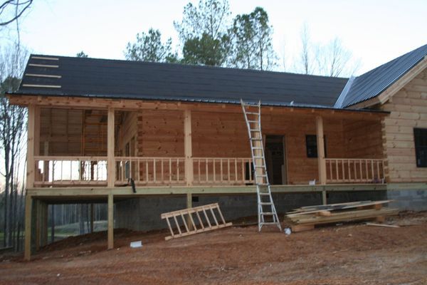 A wooden house under construction with a deck and ladder; roof partially installed, on a dirt foundation.