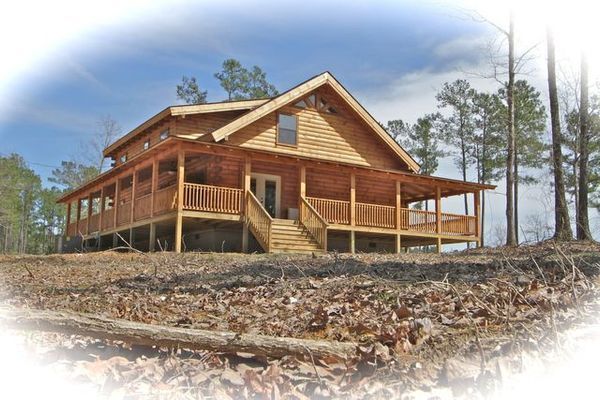 Wooden cabin with porch on a hill, surrounded by trees and dry leaves under a blue sky.