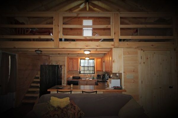 Interior of a small cabin with a loft; kitchen, living area, and stairs visible, constructed with light wood.