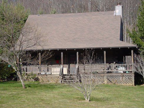 Cabin with a wraparound porch; brown roof, green grass, trees in the background.