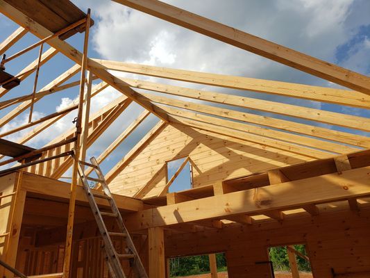 Wooden house frame under construction, scaffolding and ladder present, bright blue sky.