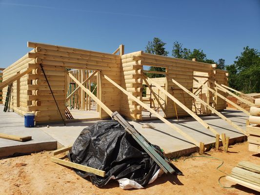 Log cabin construction site, unfinished wooden walls supported by braces on a concrete foundation, sunny day.