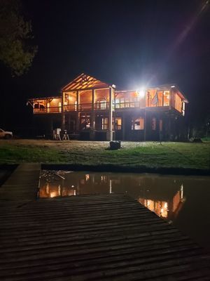 Wooden cabin lit up at night, reflecting in calm water.