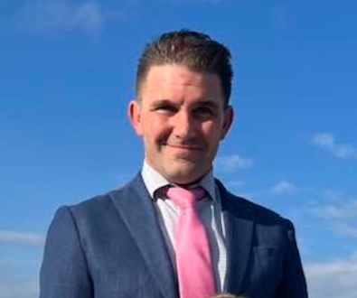 A man in a suit and pink tie is standing in front of a blue sky.