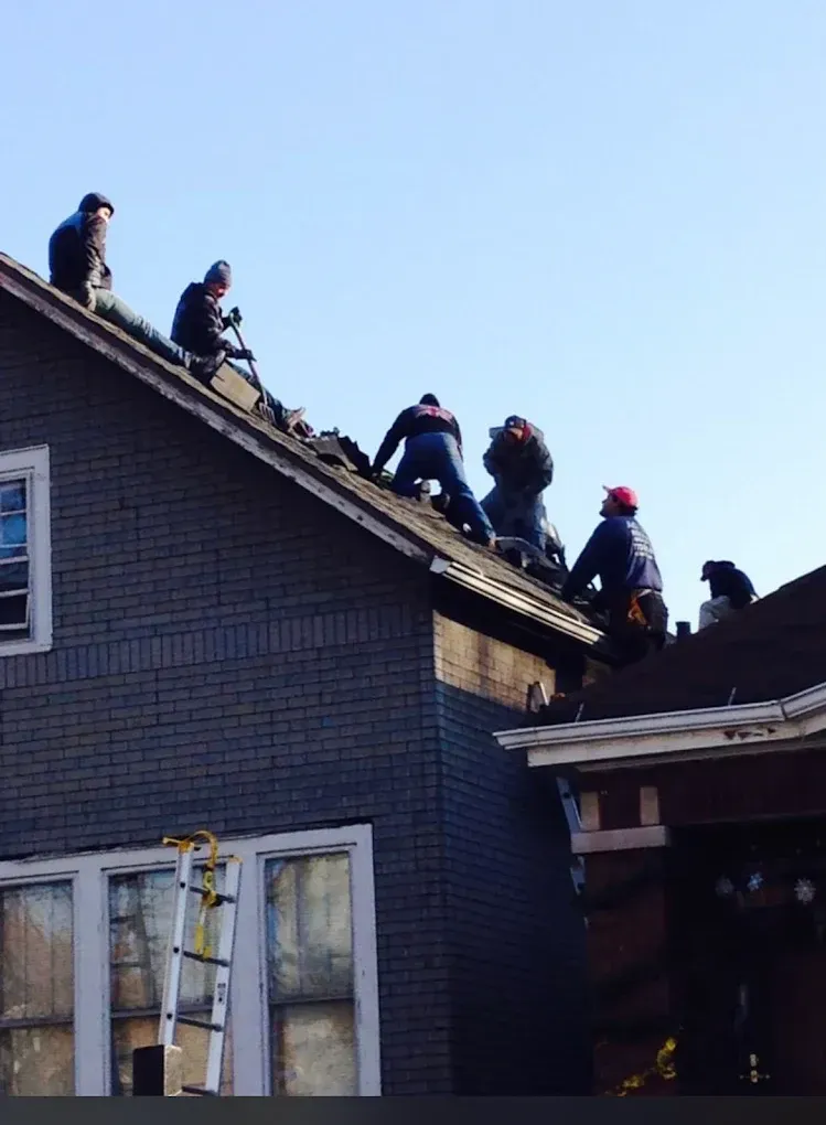 Workers on a dark roof installing shingles on a house at dusk