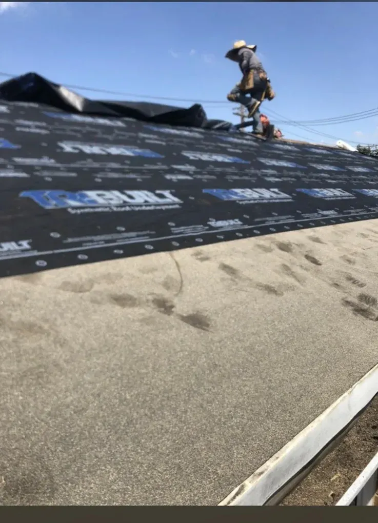 Worker installing black roofing underlayment on a sloped roof under a blue sky