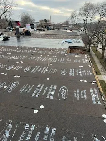 Rooftop covered with black underlayment and white printed markings, with trees and buildings in the background
