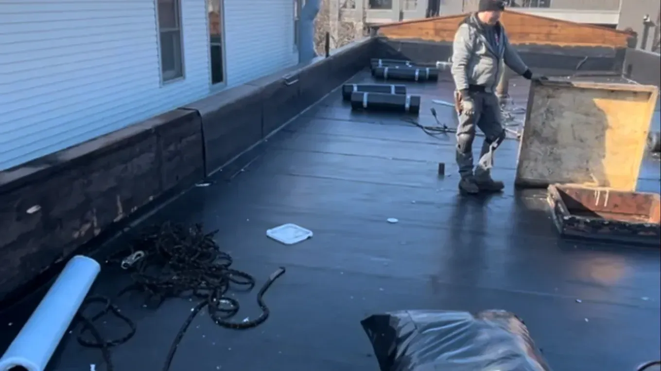 Worker repairing a flat roof beside a building, with tools and materials scattered nearby.