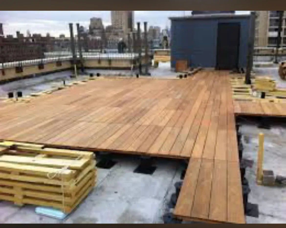 Wood deck under construction on a rooftop, with stacked lumber and a dark building entrance in the background