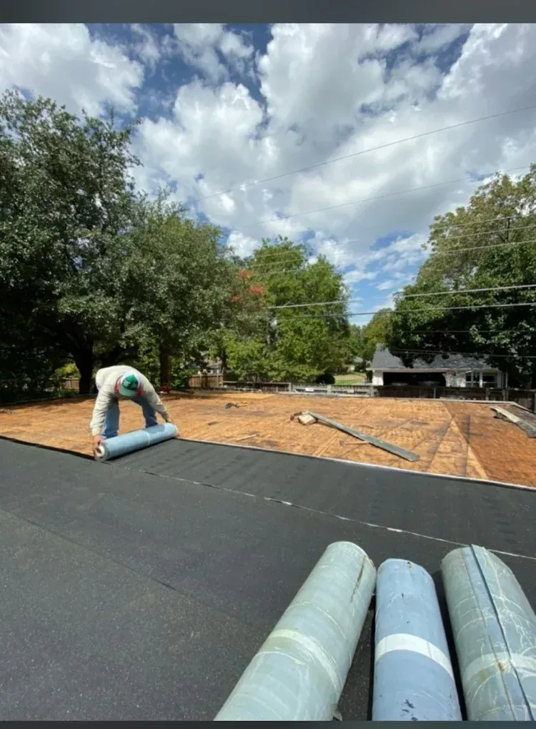 Roof with black underlayment, rolled roofing materials, and a cloudy sky over trees and houses