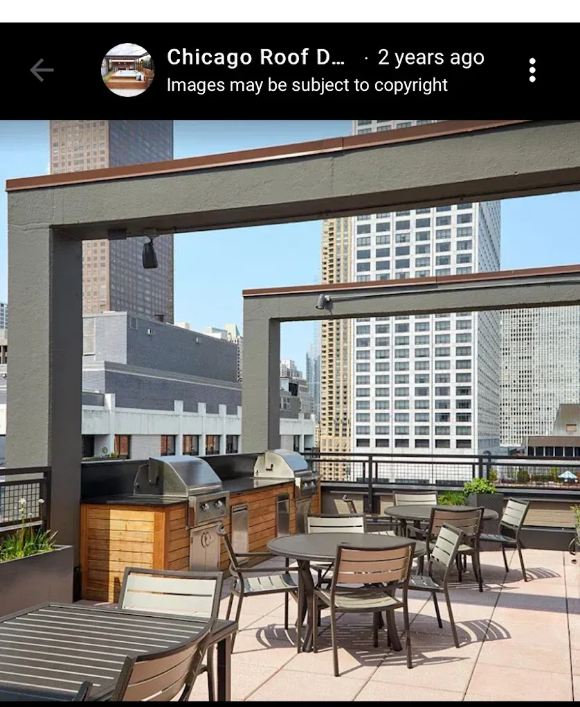 Rooftop patio with tables, chairs, and grill, overlooking city buildings under a clear sky