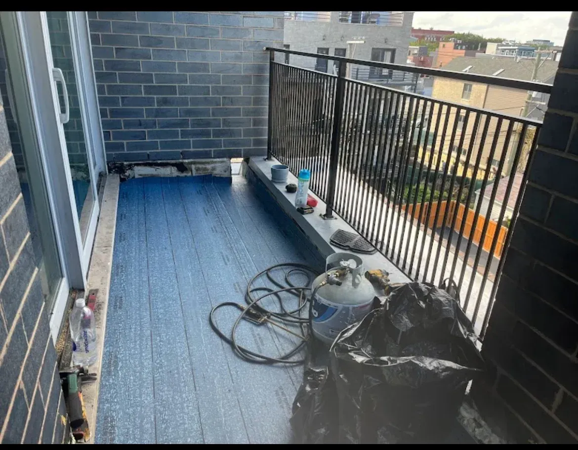 Balcony with black railing, blue floor, coiled hose, and cleaning supplies in a cluttered corner