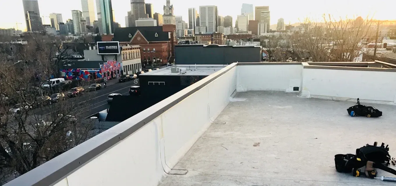 Rooftop with city skyline at sunset, white parapet walls, and two people near equipment on the right.