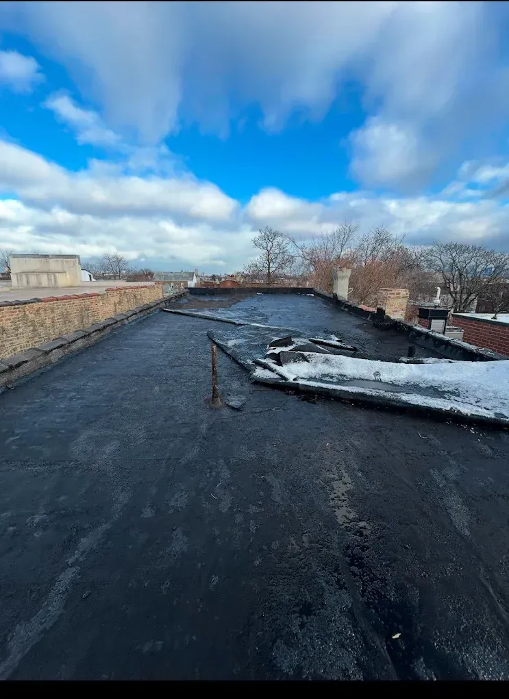 Flat black roof with wind-damaged white debris under a bright blue sky with clouds