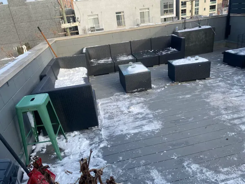 Snow-dusted rooftop patio with black wicker furniture, a green stool, and city buildings in the background