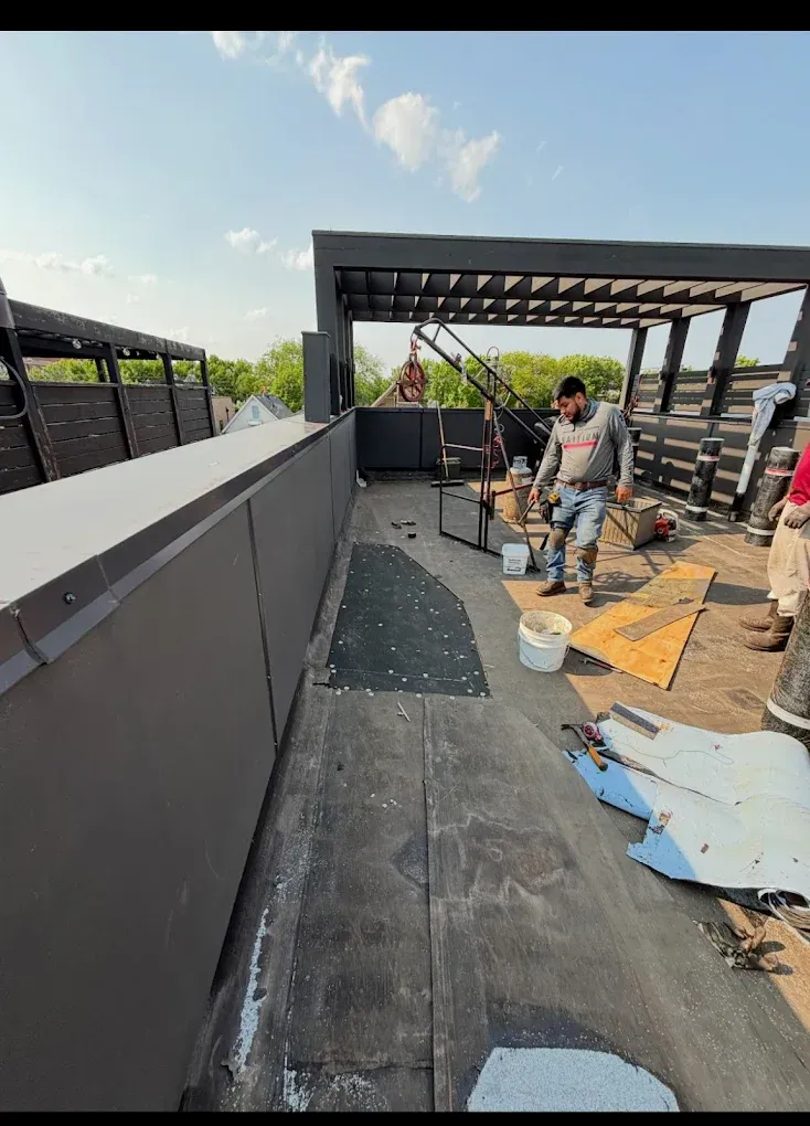 Person working on a rooftop deck beside a pergola, with tools and roofing materials scattered around.