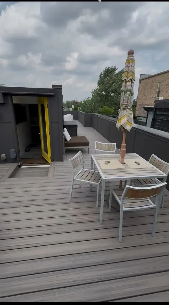 Outdoor rooftop deck with white table, chairs, closed umbrella, and cloudy sky