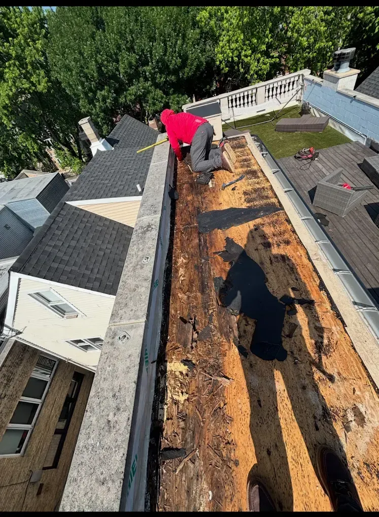 Worker on a rooftop removing old shingles from a long roof section beside a white railing.