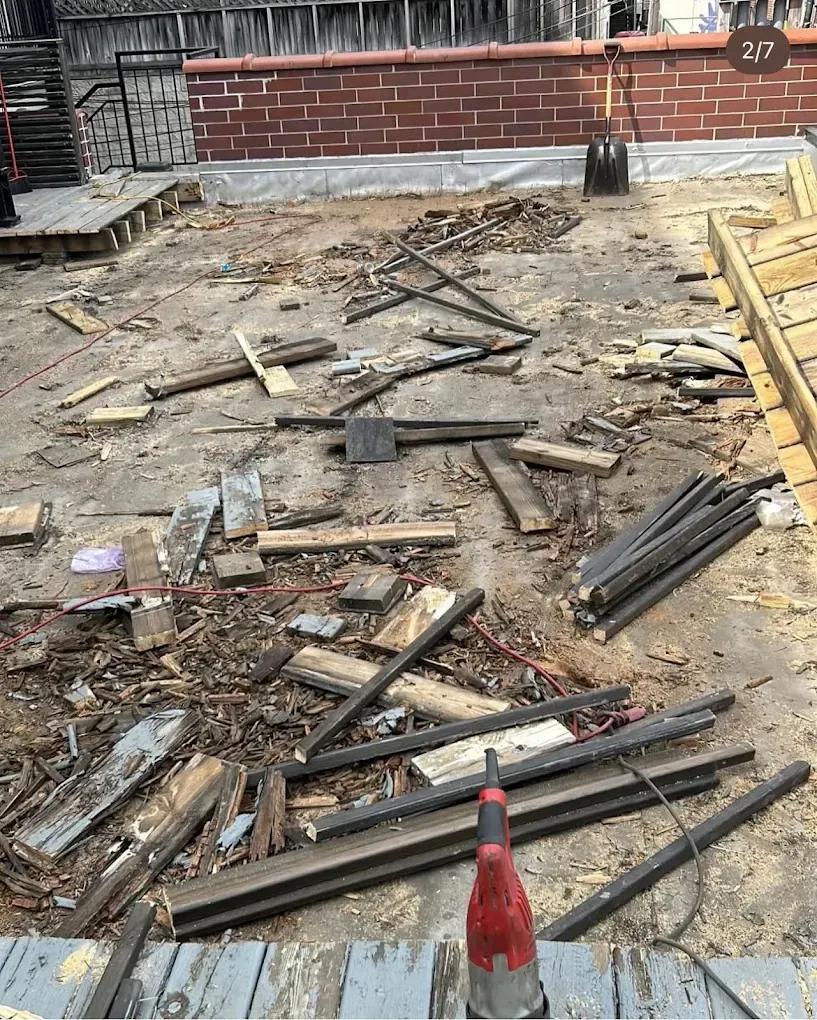 Debris and construction materials scattered on a dirt lot beside a brick wall and fence.