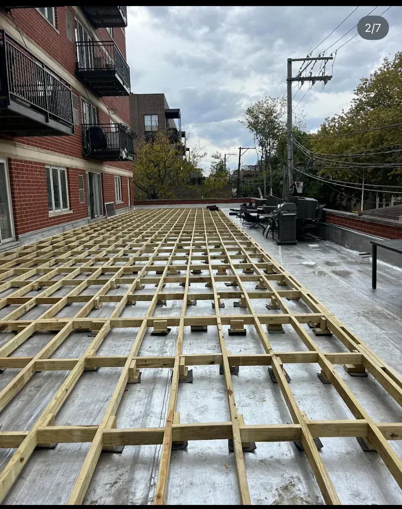 Wooden deck framing laid over a rooftop terrace beside brick buildings and trees under a cloudy sky