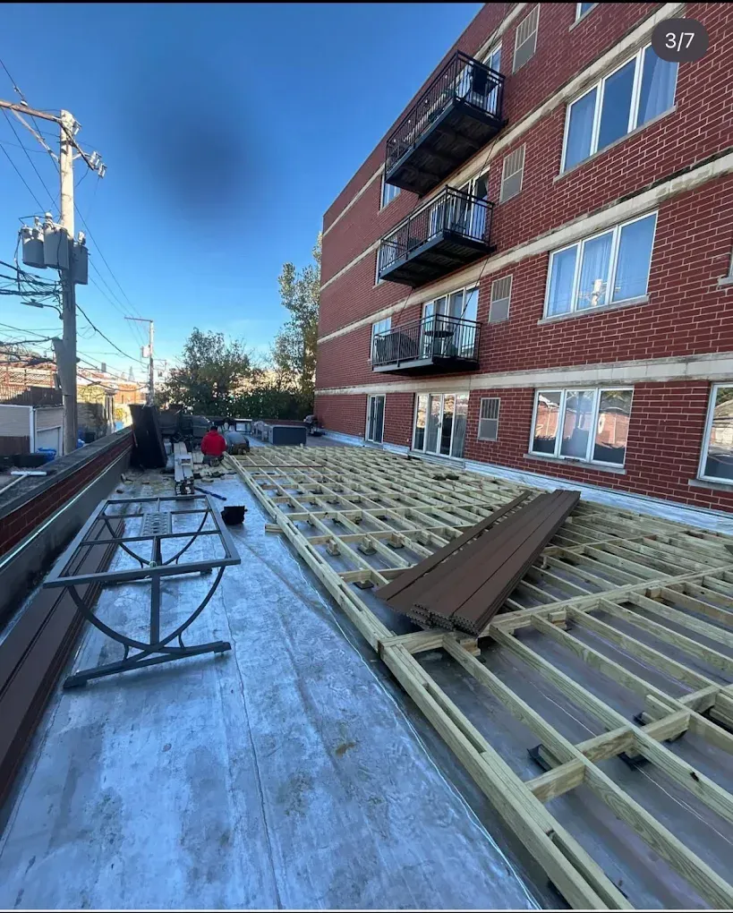 Flat rooftop under construction beside a red brick apartment building, with stacked lumber and workers in the distance.