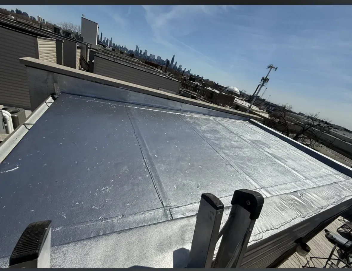 Silver reflective roof on a building under a clear blue sky, with vents and nearby rooftops visible.