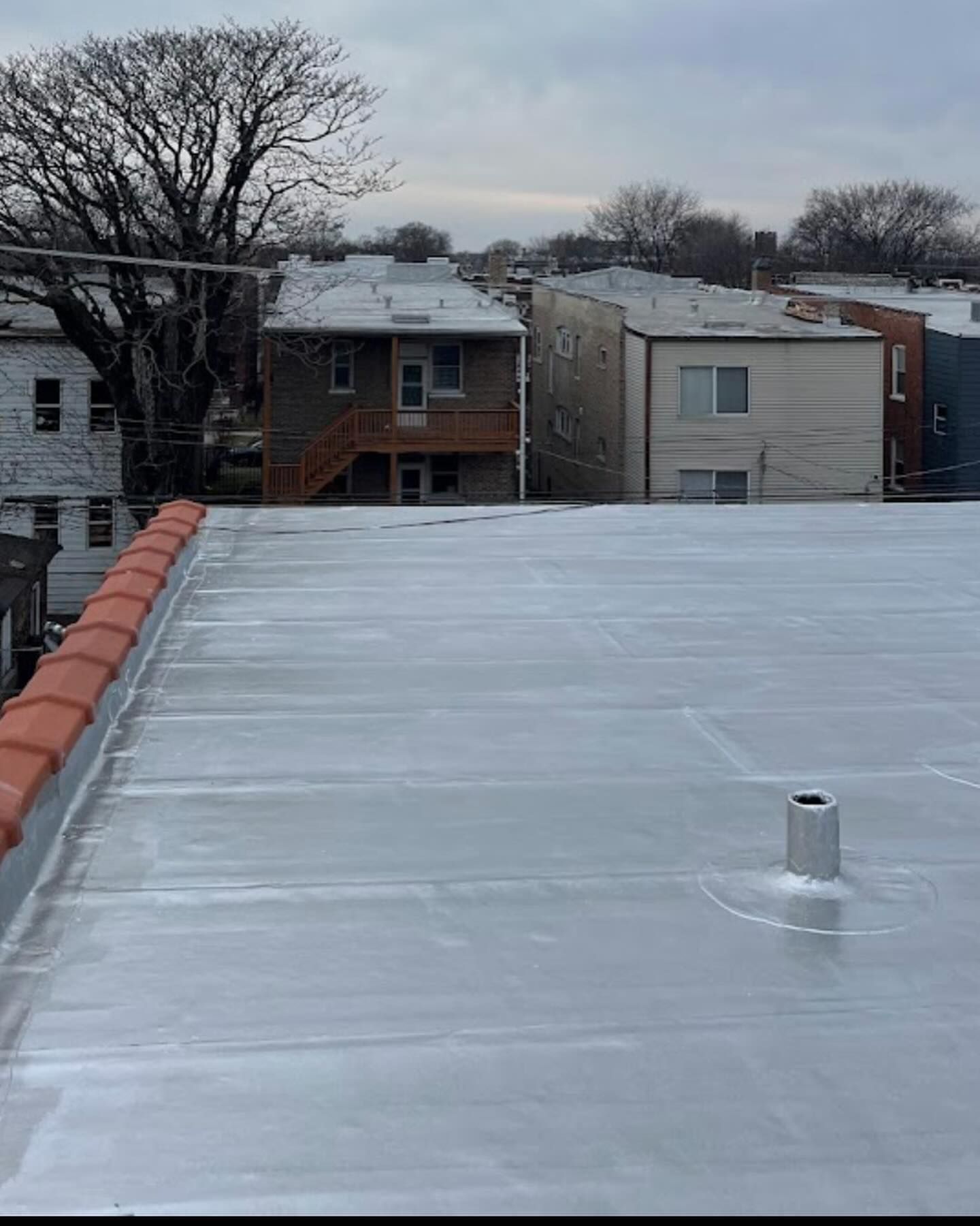 Flat white rooftop with a small vent, overlooking snowy residential buildings and bare trees under an overcast sky