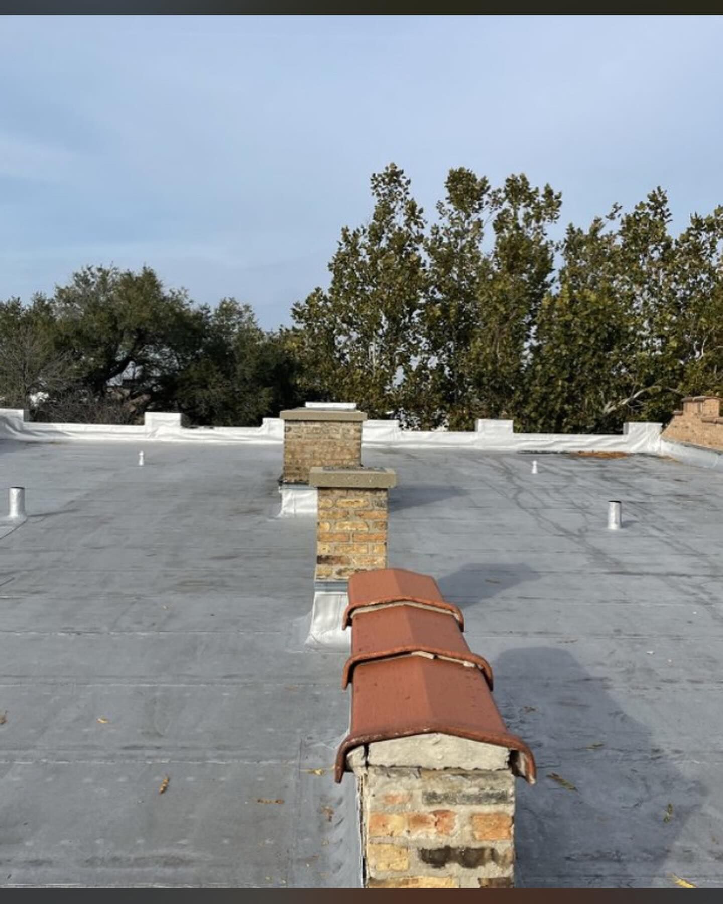 Flat rooftop with gray waterproofing, brick chimneys, and red vent covers under a blue sky.