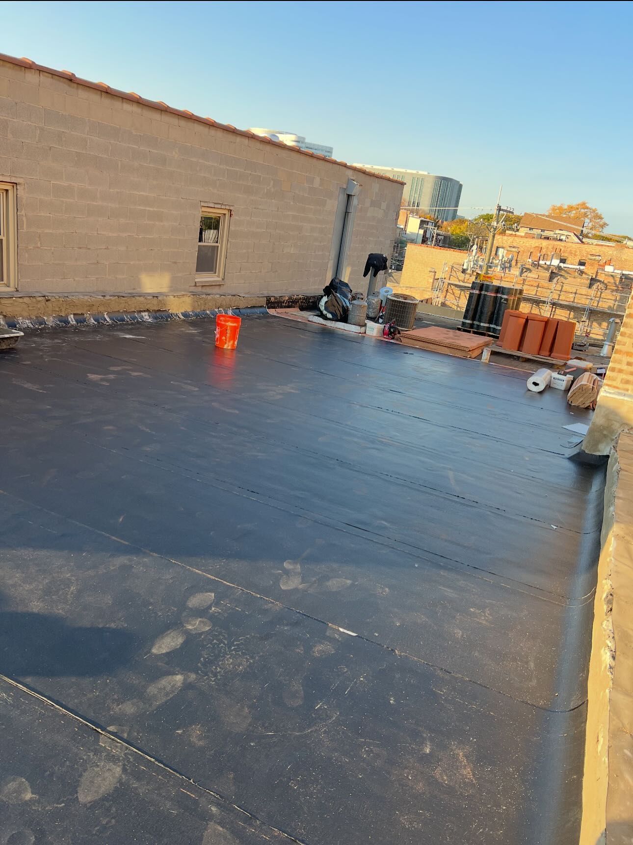 Rooftop underlayment with black roofing material, brick parapet walls, and construction tools in daylight
