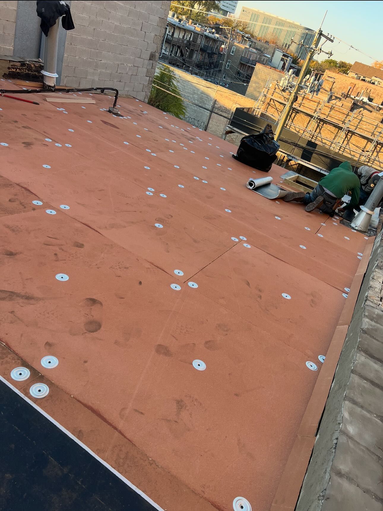 Rooftop with orange waterproof membrane, scattered fasteners, tools, and workers near the edge with city view