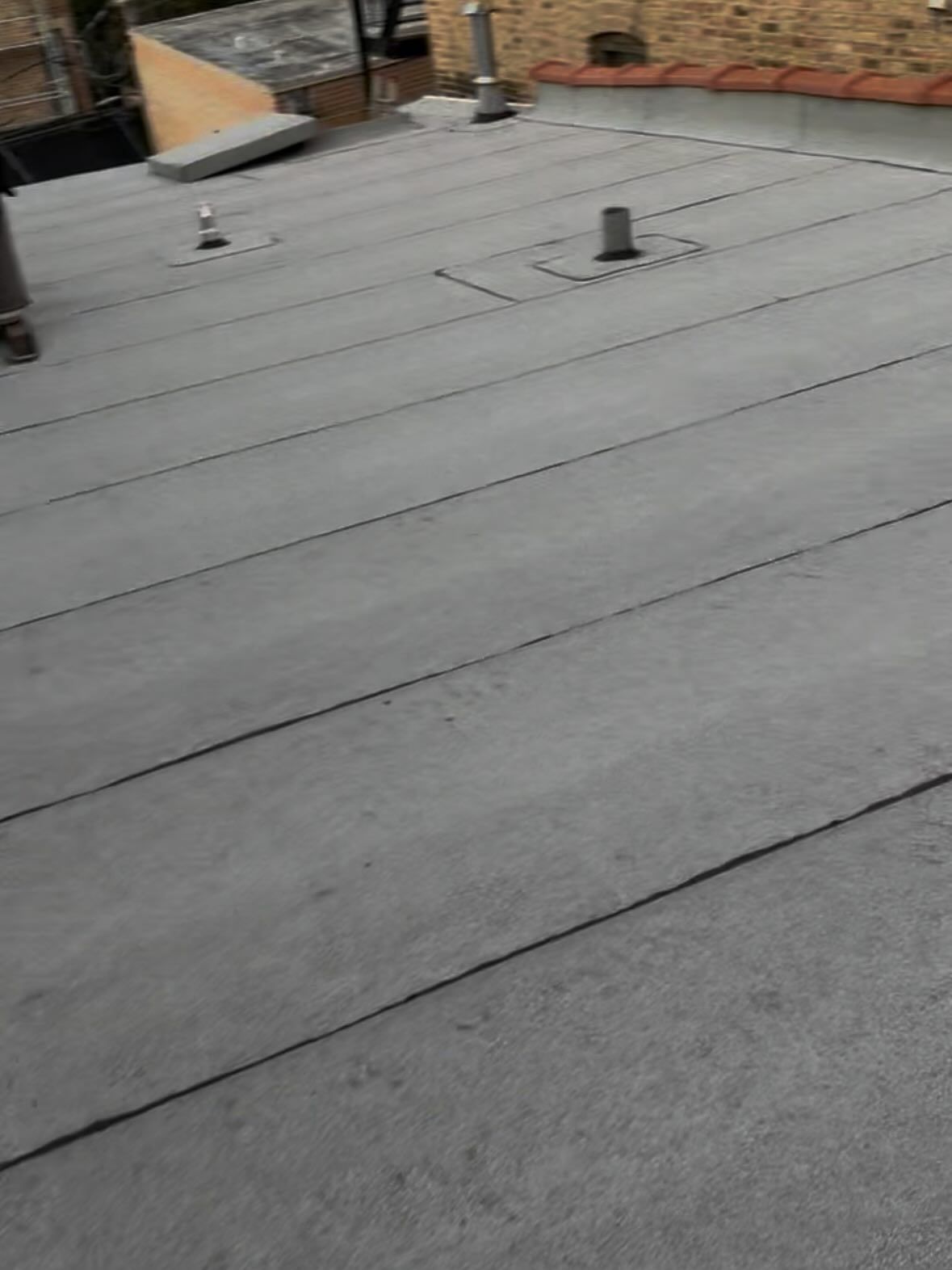Flat gray roof with seams and two vent pipes near a brick building edge