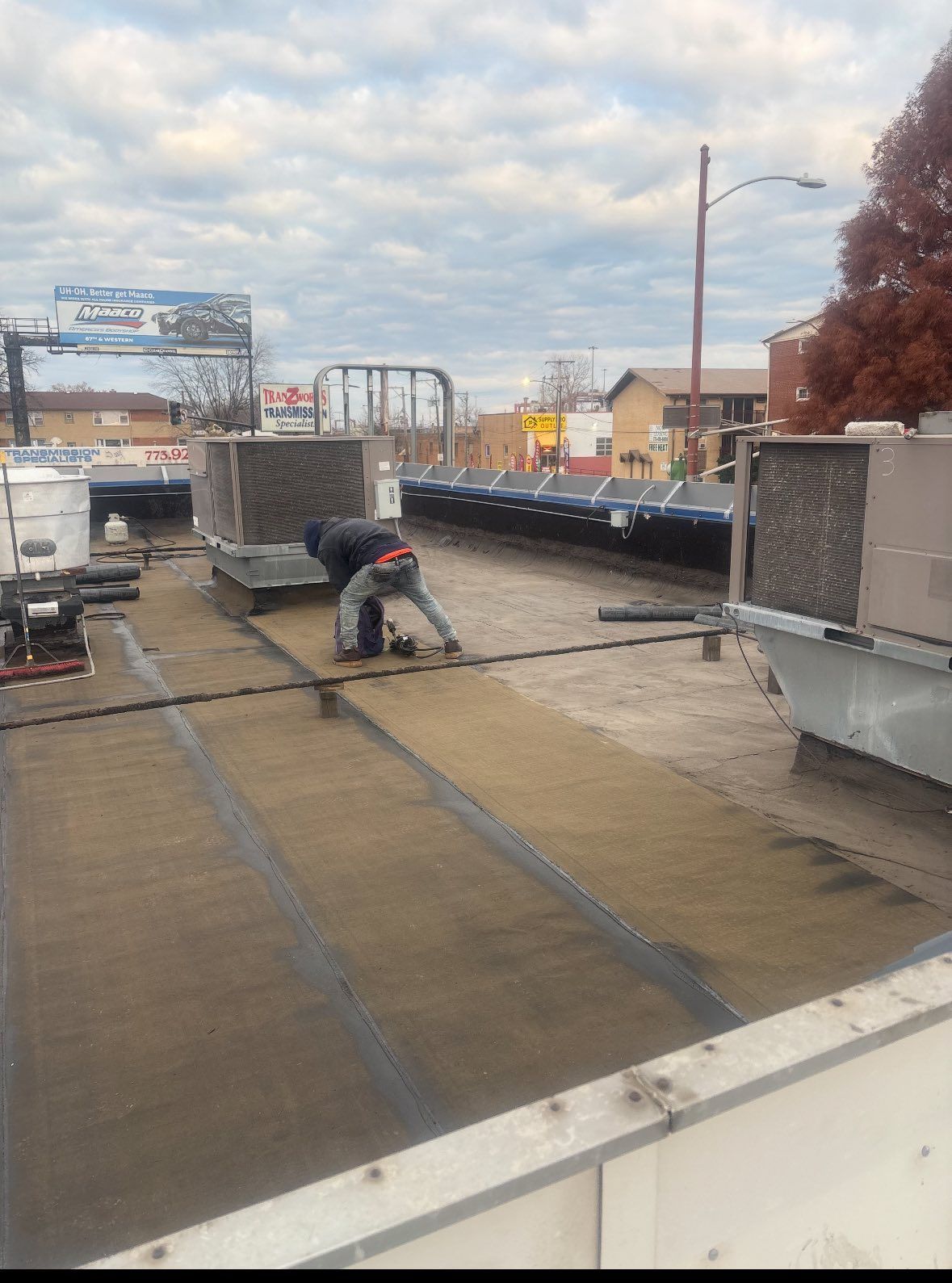 Worker on a flat rooftop near HVAC units, inspecting or repairing the roof under a cloudy sky.