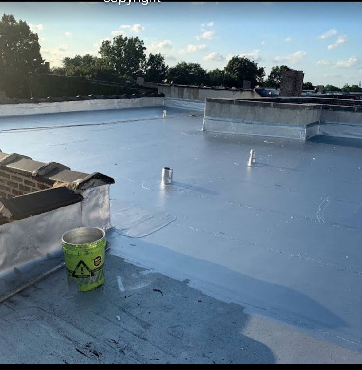 Flat white rooftop with blue membrane, vents, and a green bucket in the foreground under a clear sky