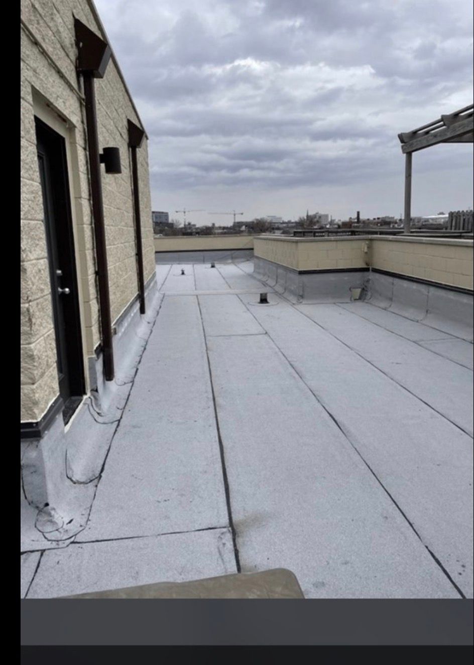 Rooftop with gray membrane roofing beside a brick wall under a cloudy sky.