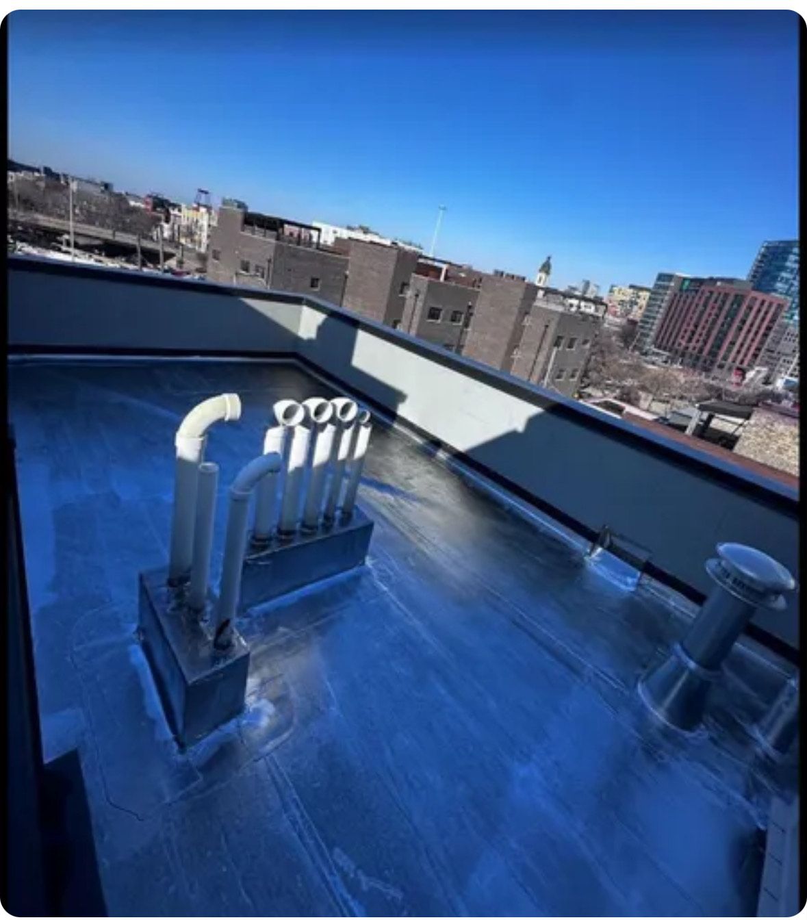 Rooftop ventilation pipes and ductwork on a reflective metal roof with city buildings in the background.