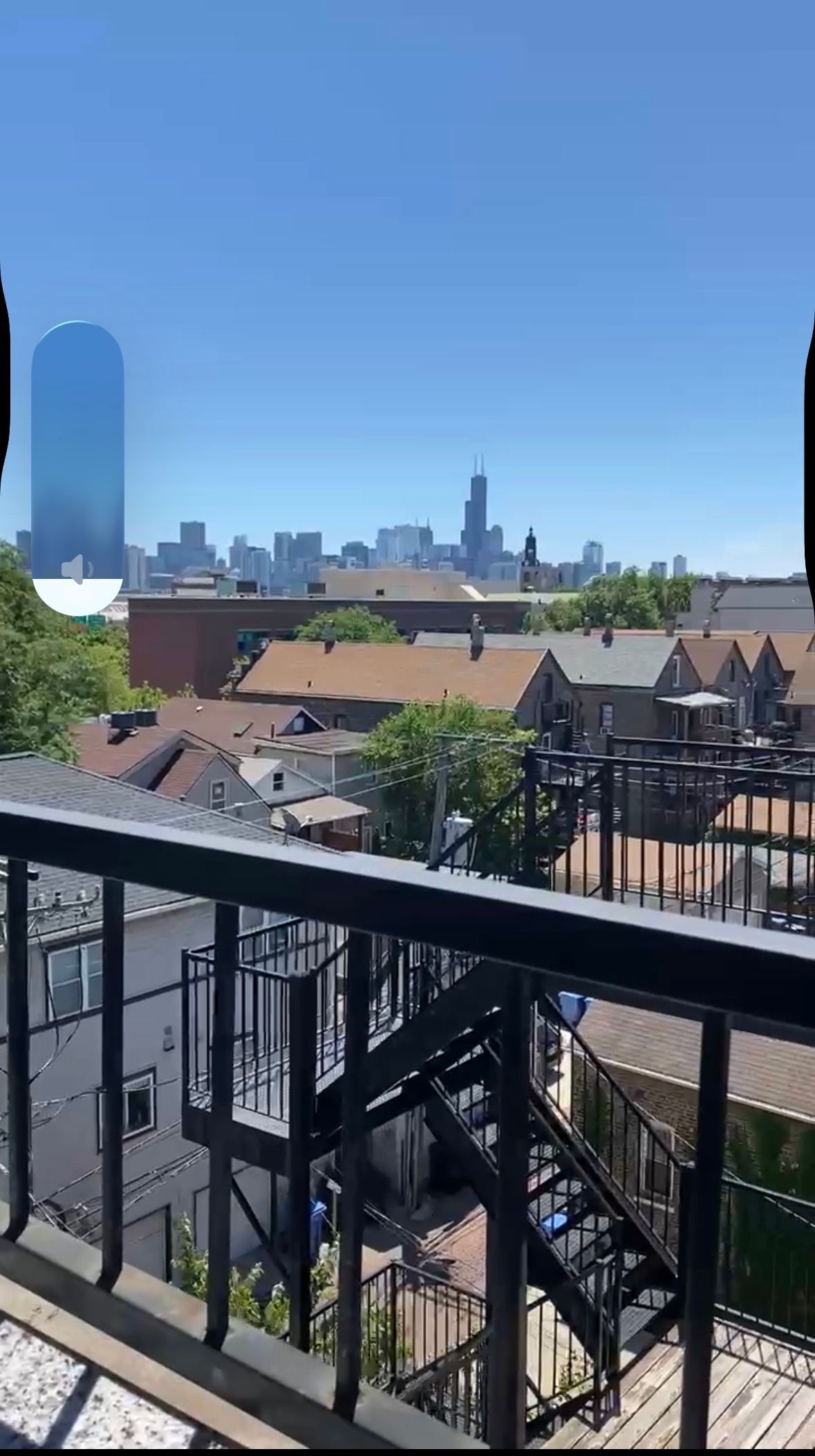 City skyline view from a balcony with rooftops, a blue sky, and black railing in the foreground