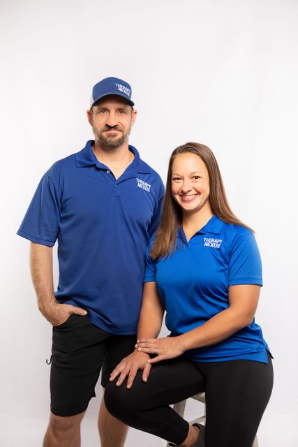 Two people in blue shirts, smiling, posing in front of a white backdrop. Man in hat. Woman sits.