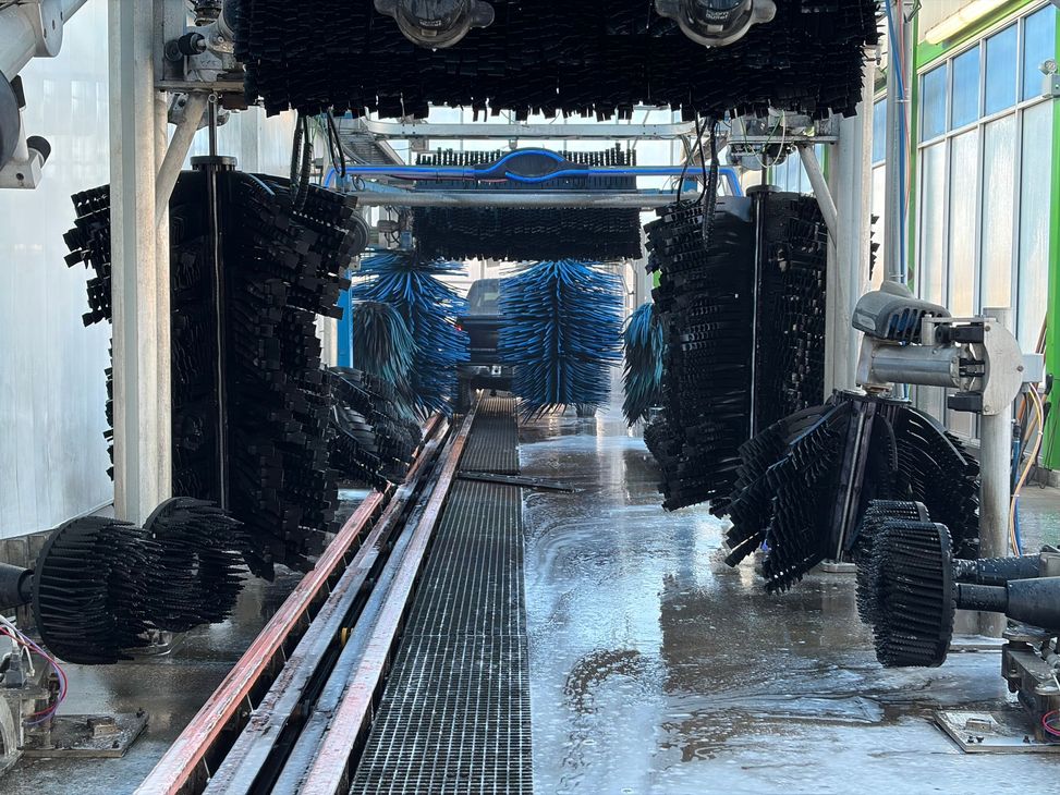 Car wash tunnel with spinning black brushes, wet floor, and cars being cleaned inside.