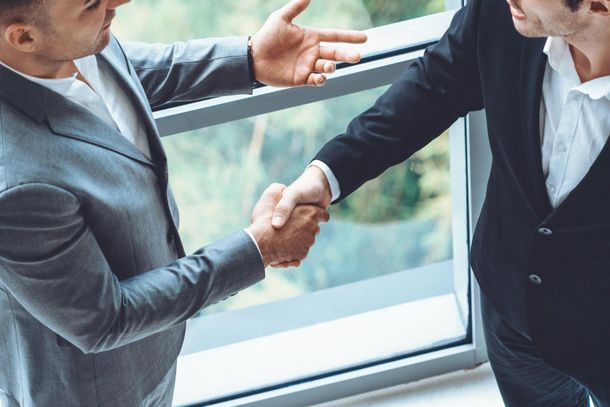 Two men in suits shaking hands, a gesture of agreement, by a window.