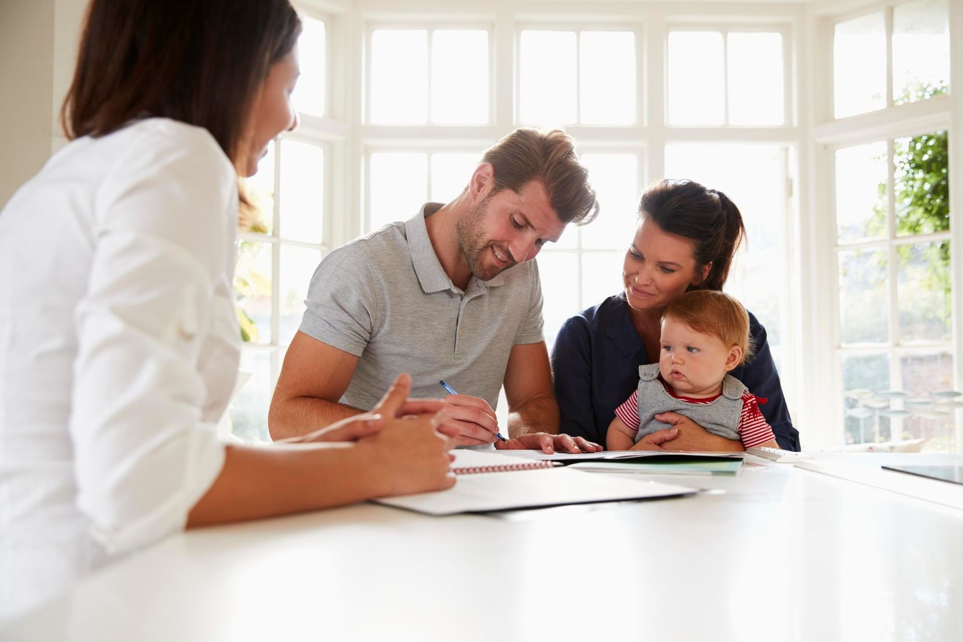 A woman advising a family, including a baby, at a table in a sunlit room. They are reviewing documents.