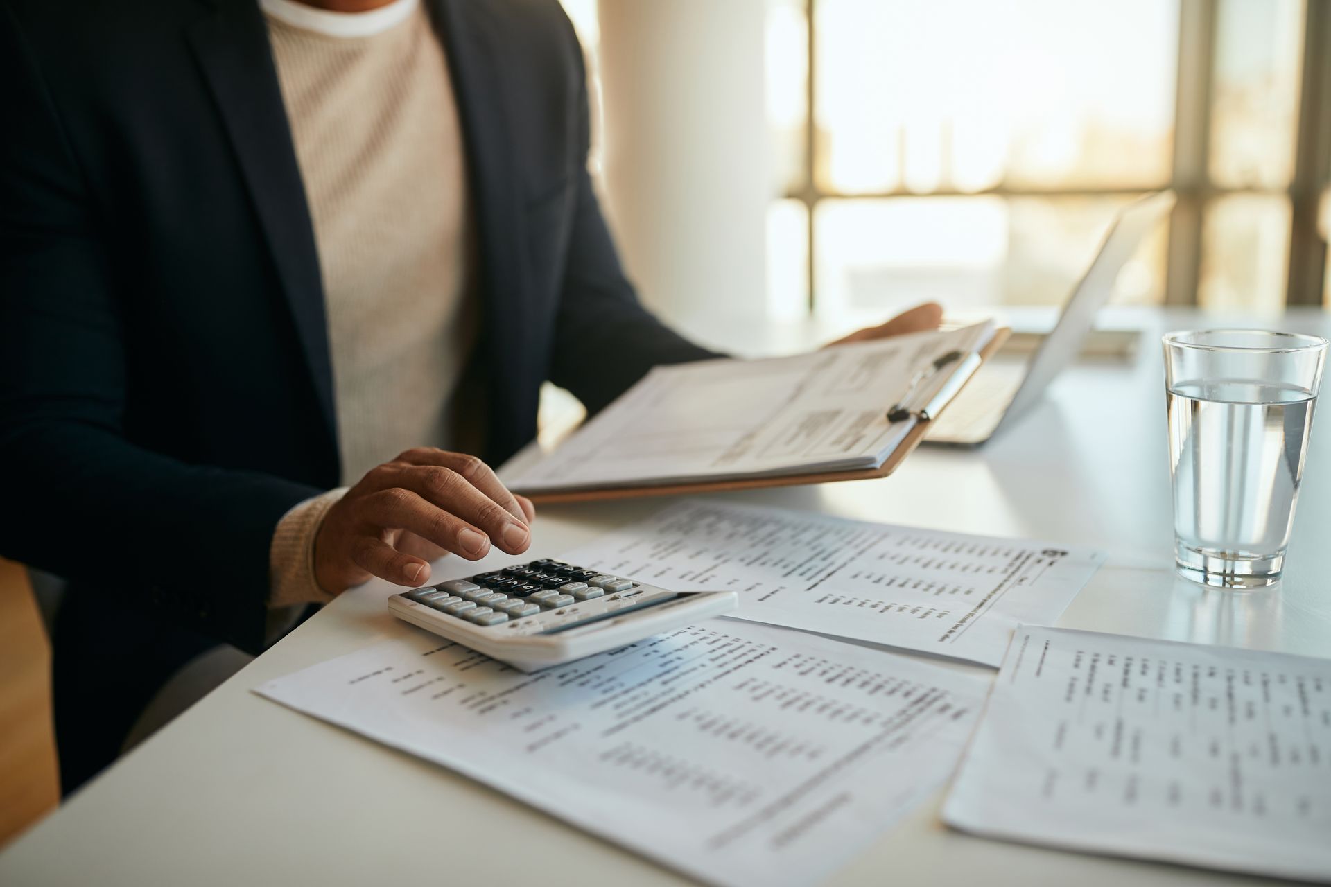 Person in blazer using calculator, reviewing paperwork, and near a laptop and a glass of water.
